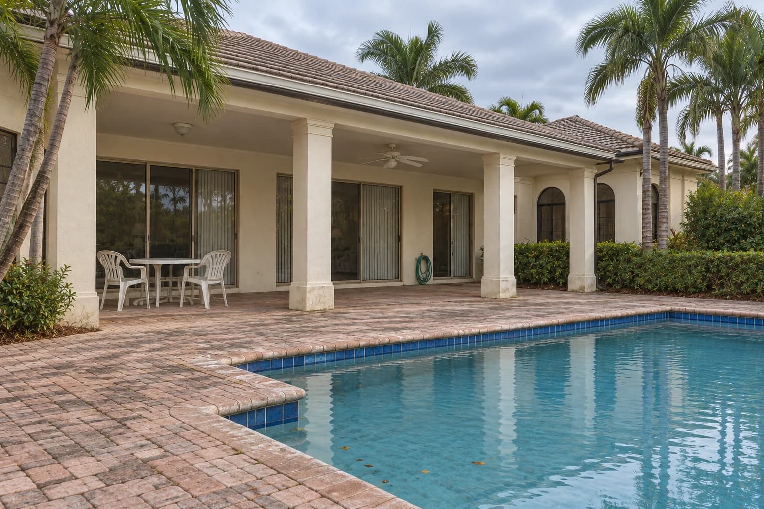 The Palmetto Terrace — Before concept: a modest Naples pool terrace under cloudy skies with weathered paving, older pool finishes, a plain covered patio, and basic plastic furniture